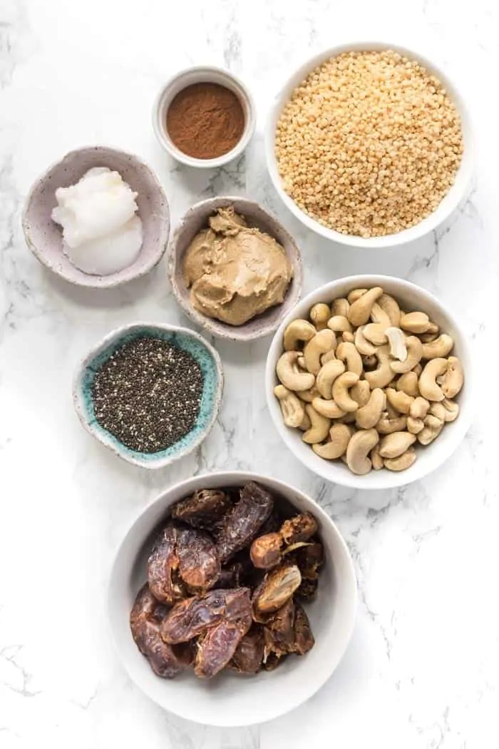 Overhead view of a bowl of dates, a bowl of cashews, a bowl of peanut butter, a bowl of chia seeds, a bowl of puffed quinoa, a bowl of coconut oil, and a bowl of cinnamon. 
