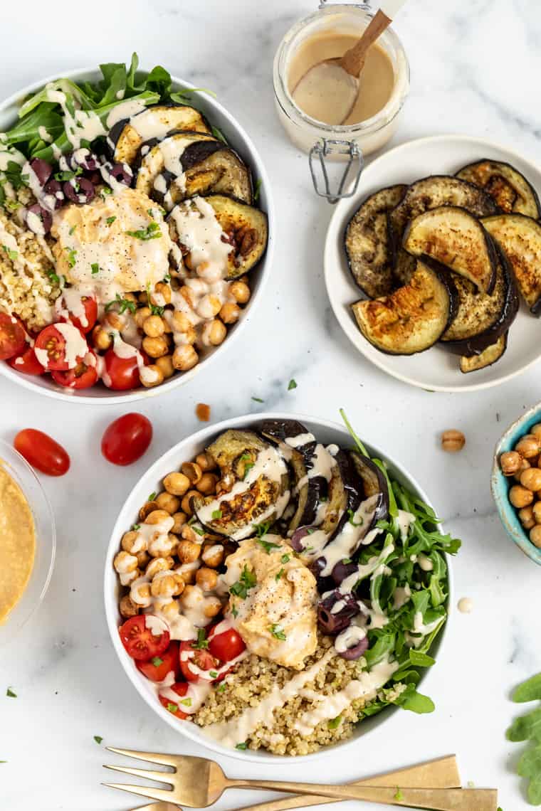 Overhead view of two quinoa bowls topped with tomatoes, olives, arugula, hummus, eggplant, chickpeas, and tahini dressing ,next to a plate of roasted eggplant, a bowl of chickpeas, some cherry tomatoes, and a jar of dressing