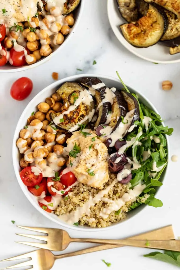 These easy Mediterranean quinoa bowls have roasted eggplant, crispy chickpeas, and hummus. They're super healthy, vegan, and gluten-free! Overhead view of a quinoa bowl topped with hummus, arugula, olives, tomatoes, chickpeas, and eggplant, next to two forks
