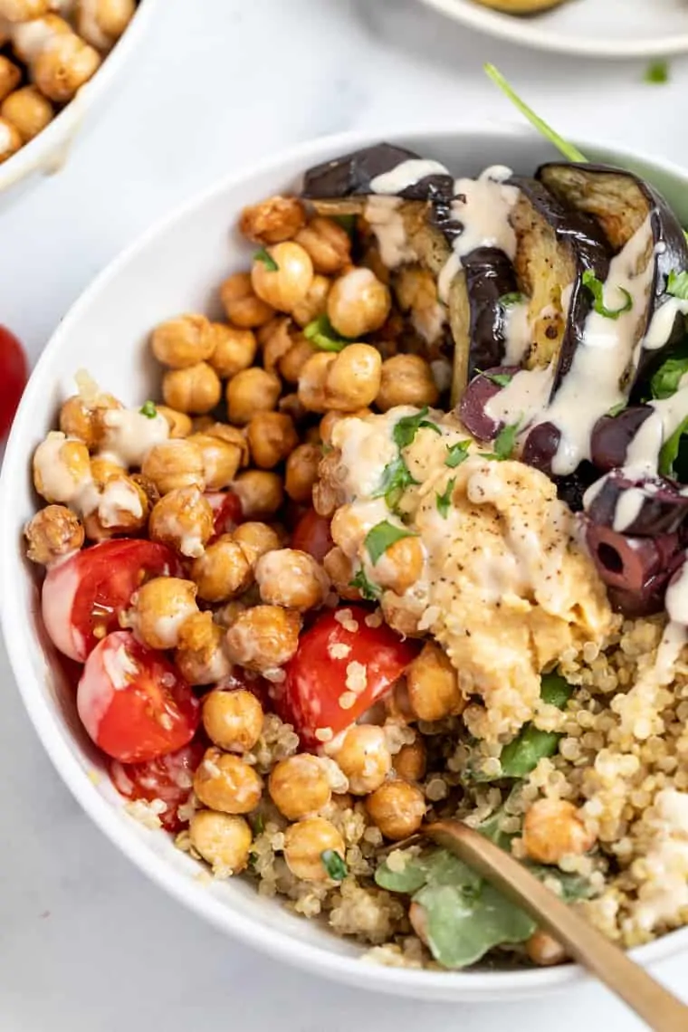 Close up of a quinoa bowl topped with eggplant, tomatoes, olives, chickpeas, and tahini dressing, with a fork in it 