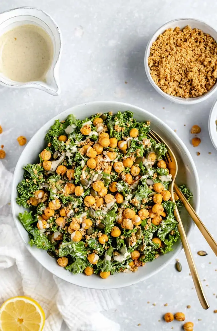 Overhead view of a bowl of kale Caesar salad topped with chickpeas, with a fork and spoon in the bowl, next to a bowl of dressing and a bowl of quinoa crispies