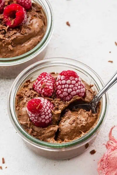 A jar of chocolate avocado mousse, topped with raspberries, with a spoon in it
