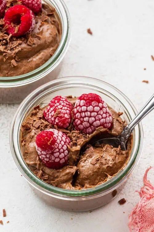 A jar of chocolate avocado mousse, topped with raspberries, with a spoon in it