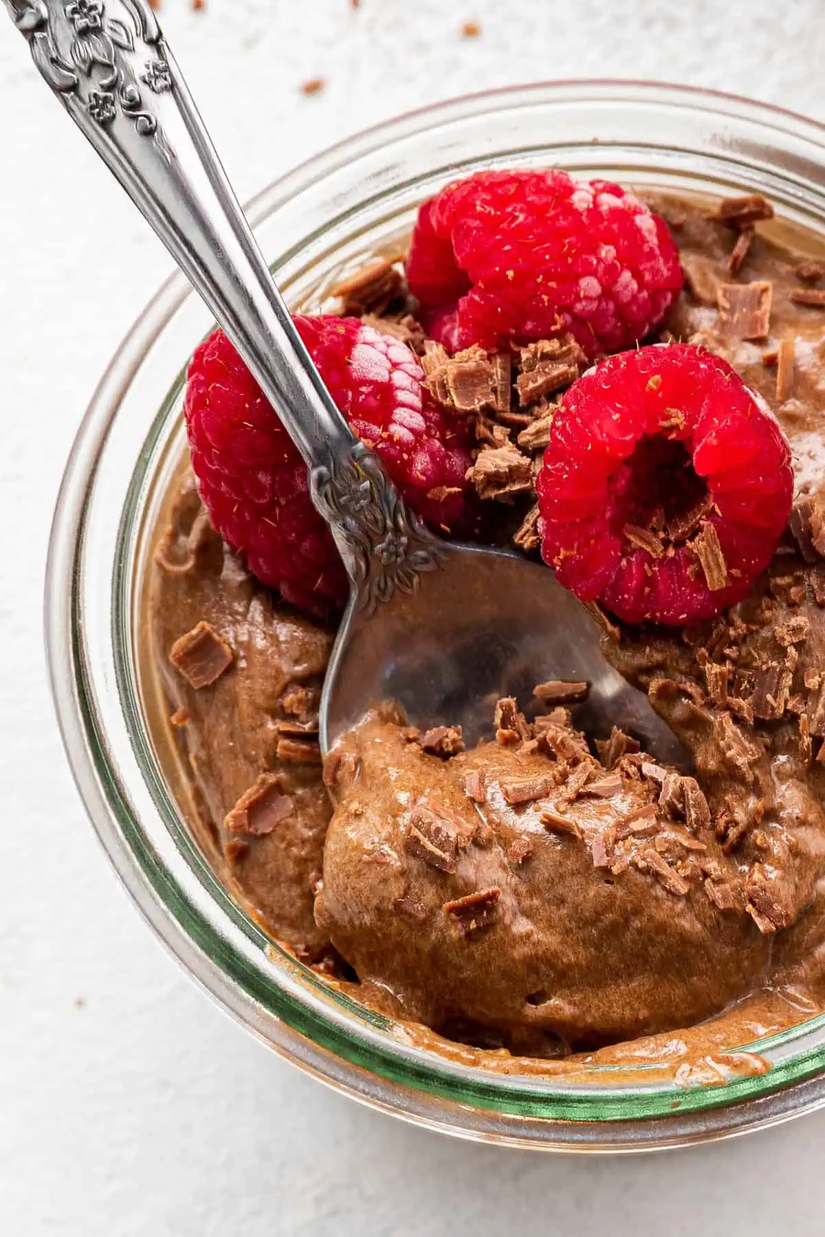 A spoon dipping into a cup of chocolate avocado mousse topped with raspberries
