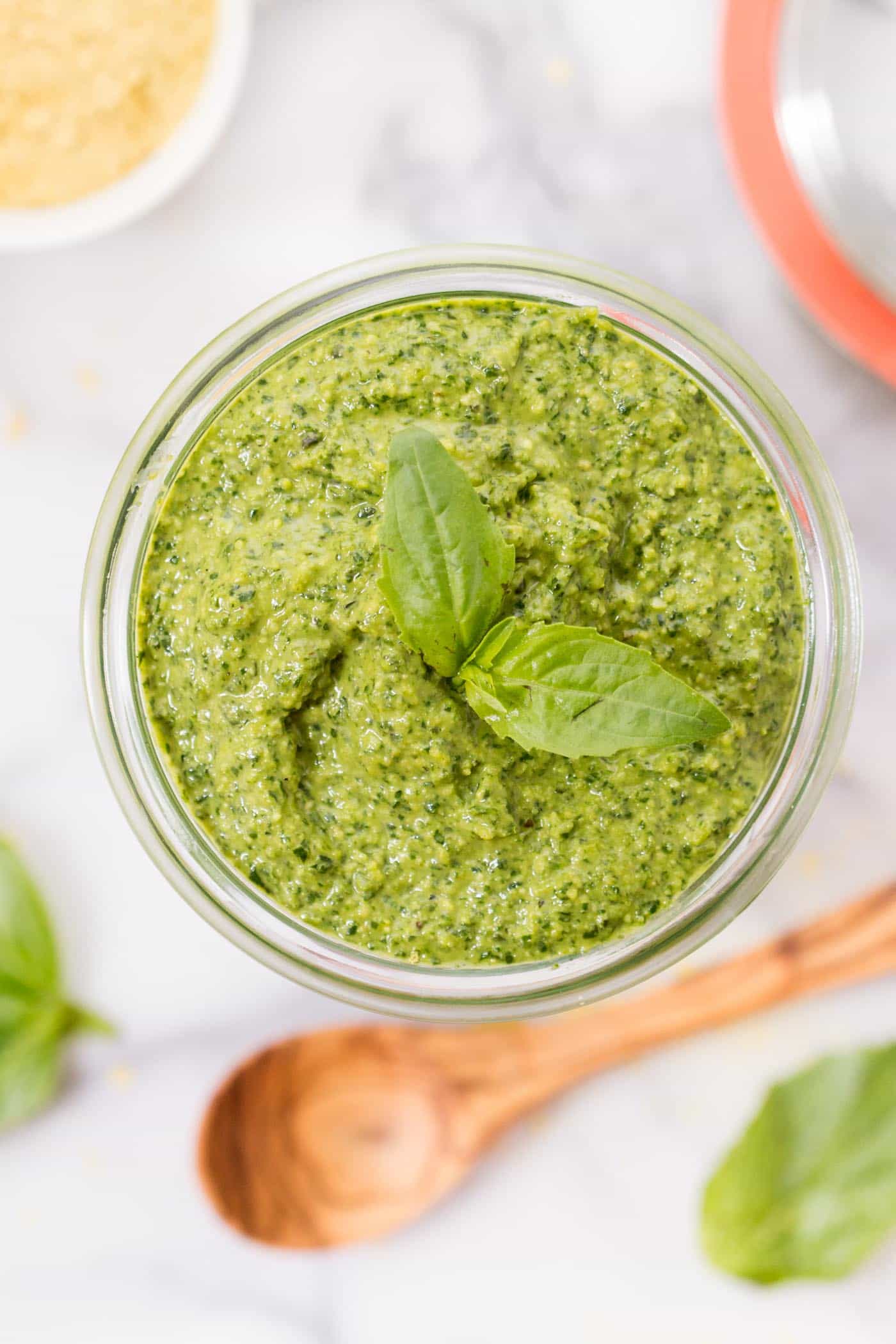 Overhead view of a jar of pesto with a sprig of basil on top, with a wooden spoon and basil leaves to the side