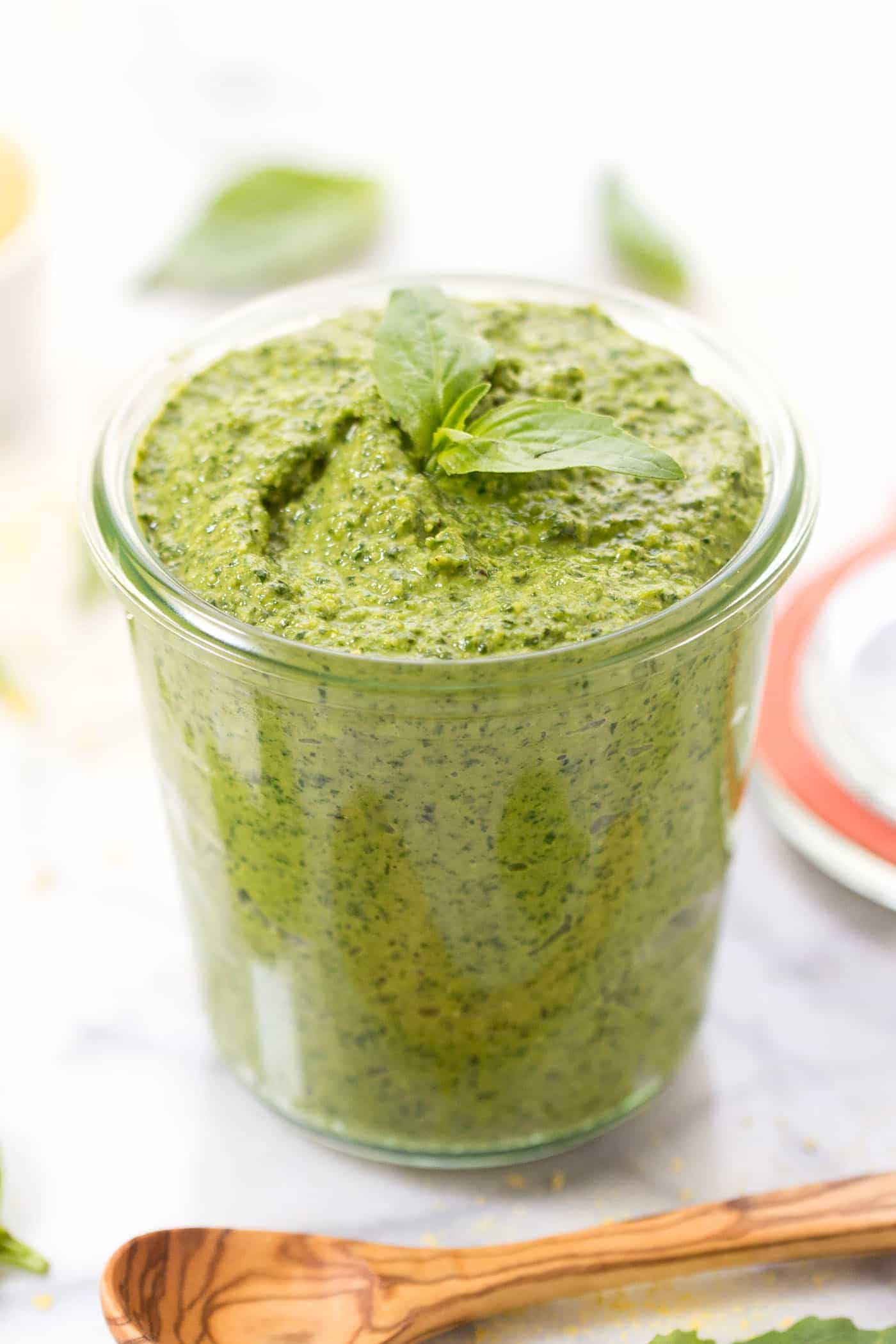 A jar full of pesto, with a sprig of fresh basil on top, and a wooden spoon in front of the jar