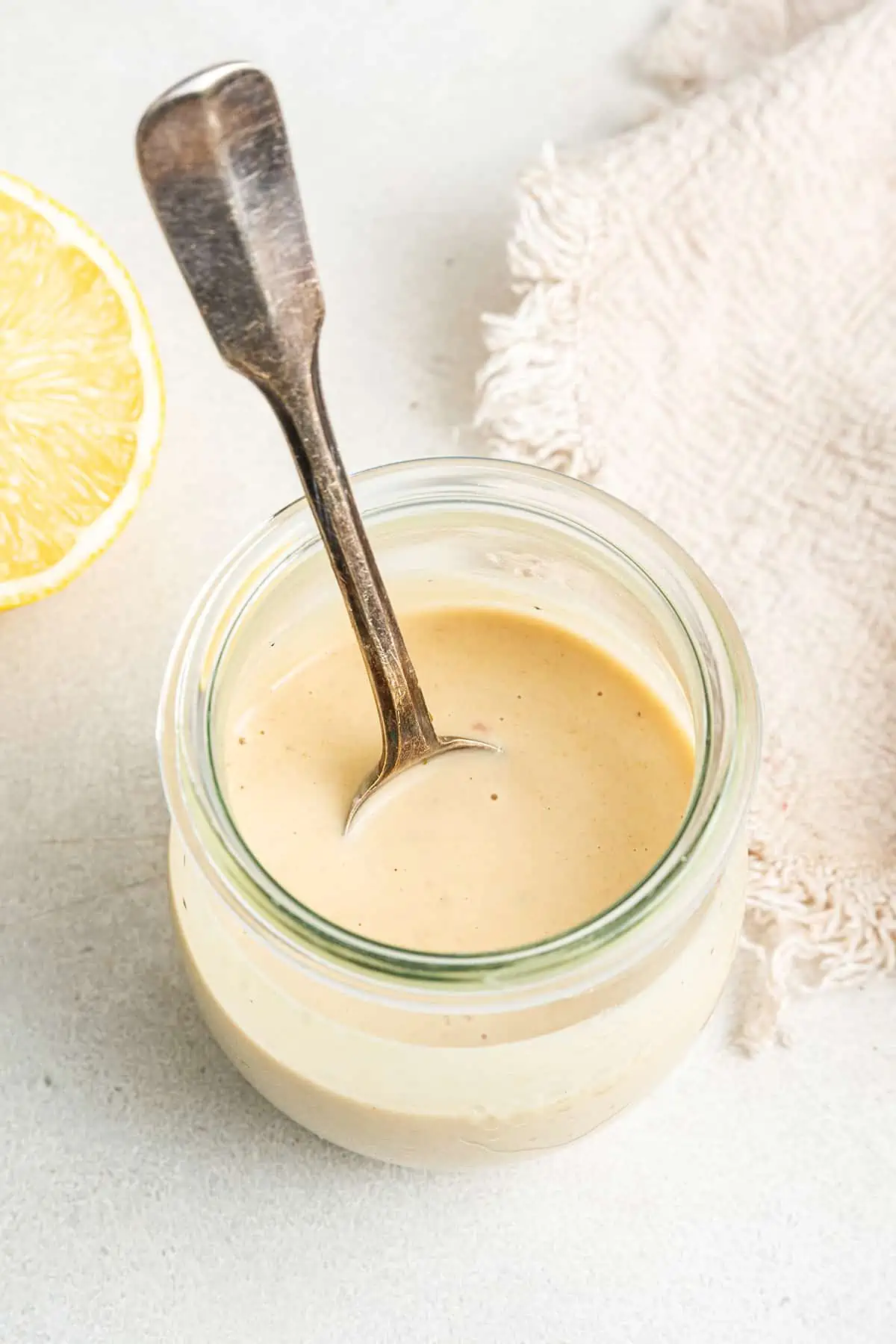 A jar of tahini dressing with a spoon in it, next to a lemon half