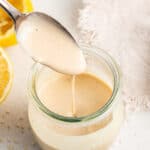 A spoon being pulled out of a jar of tahini dressing, next to some lemon halves