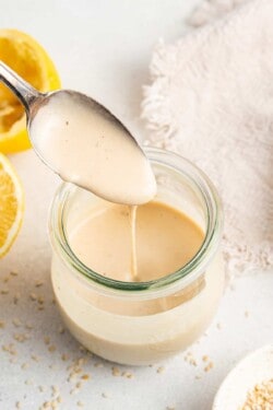 A spoon being pulled out of a jar of tahini dressing, next to some lemon halves