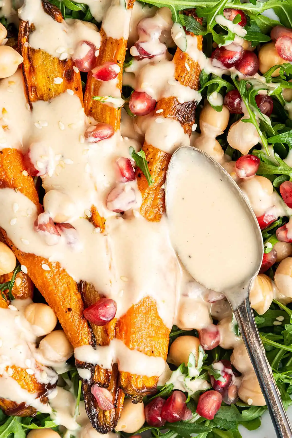 Close up of a spoon pouring tahini dressing on top of roast carrots, on top of arugula, chickpeas, and pomegranate seeds
