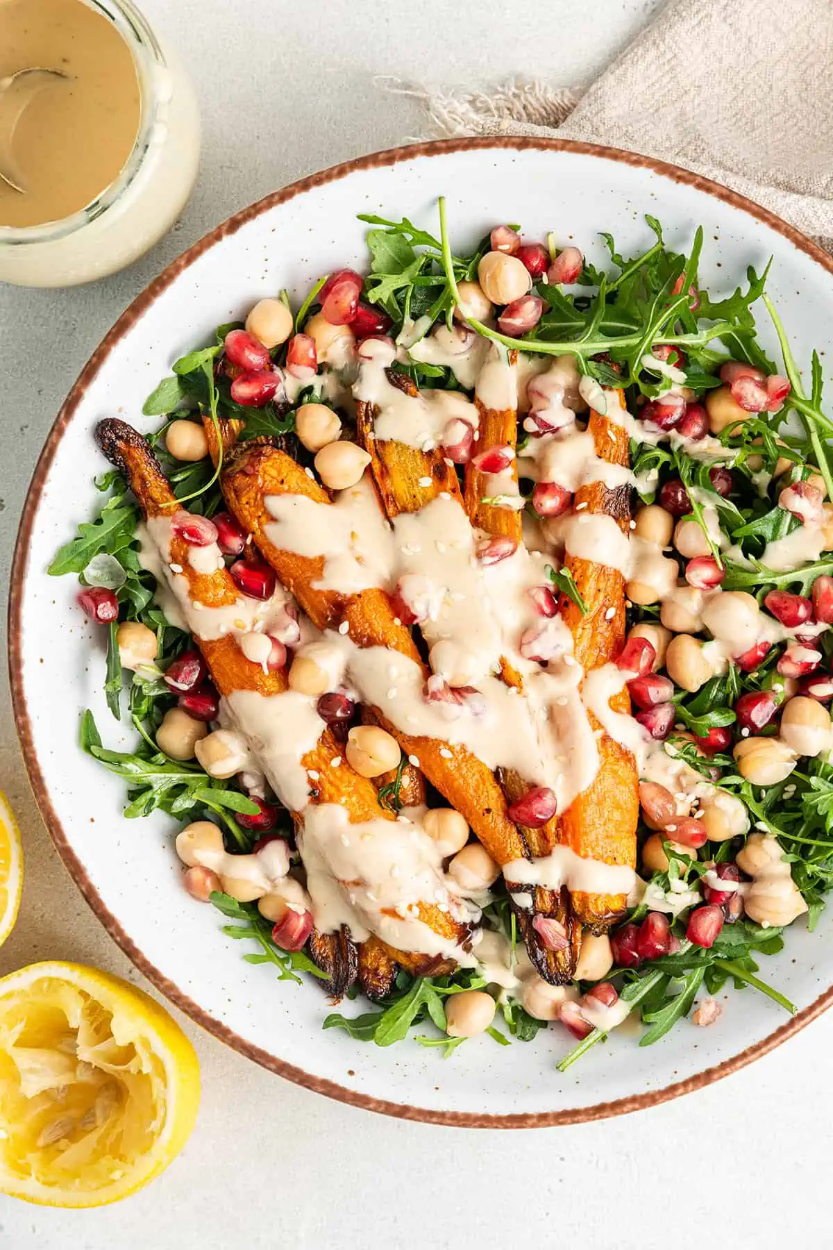 Overhead view of a salad with arugula, chickpeas, pomegranate seeds, and roasted carrots, with tahini dressing on top