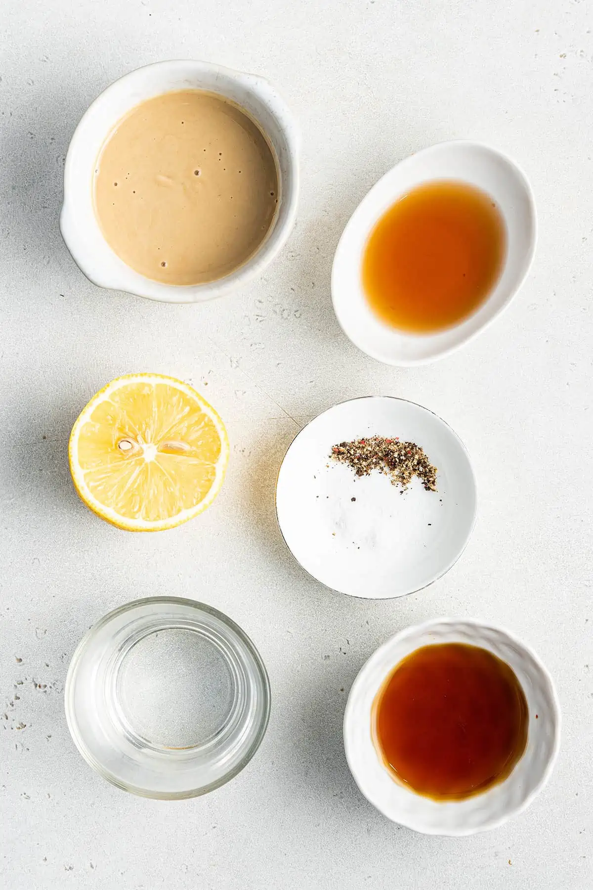 Overhead view of the ingredients needed for maple tahini dressing: half a lemon, and bowls of tahini, apple cider vinegar, maple syrup, water, and salt and pepper