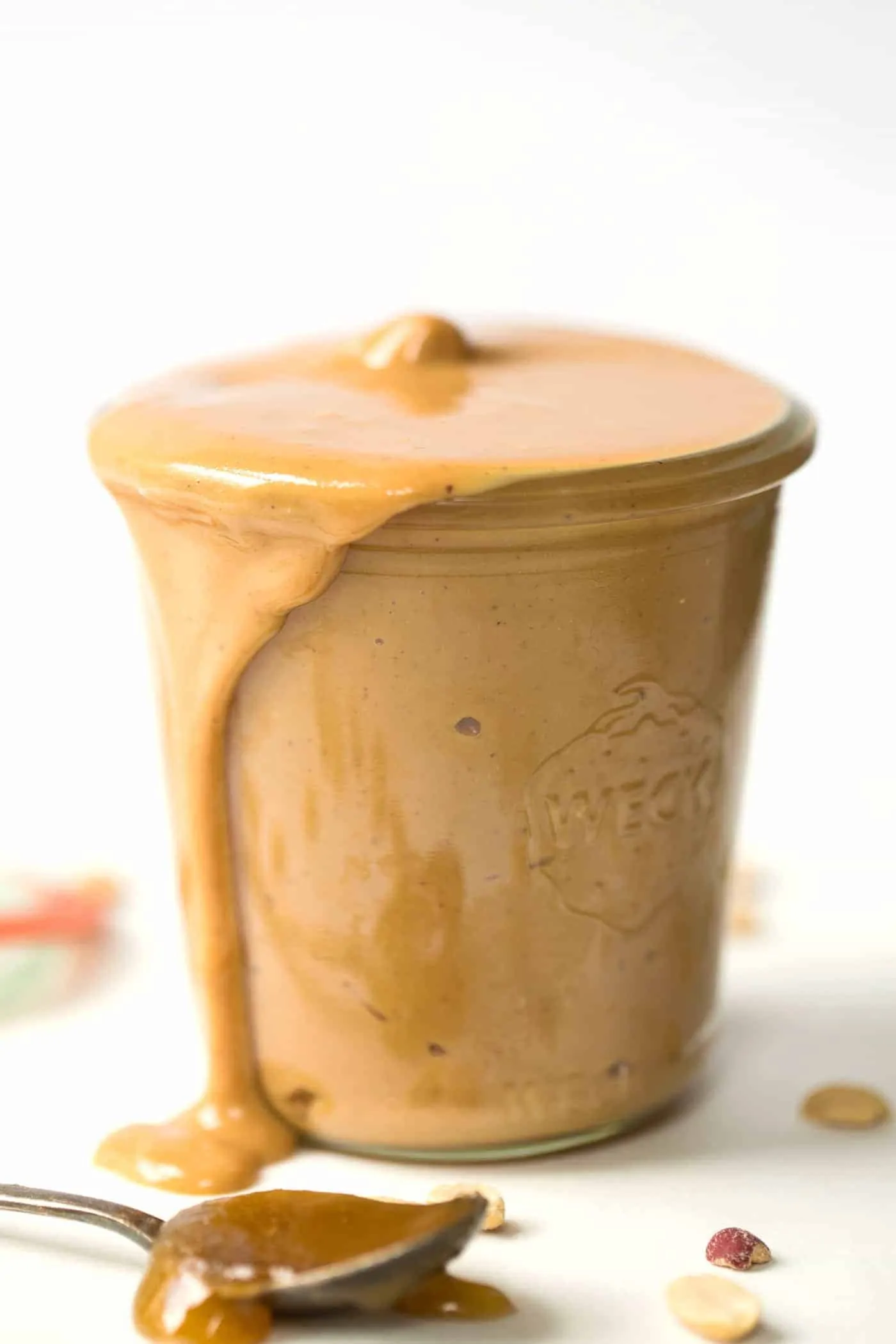 A jar of homemade peanut butter with the peanut butter dripping out, next to peanuts and a spoon covered in honey