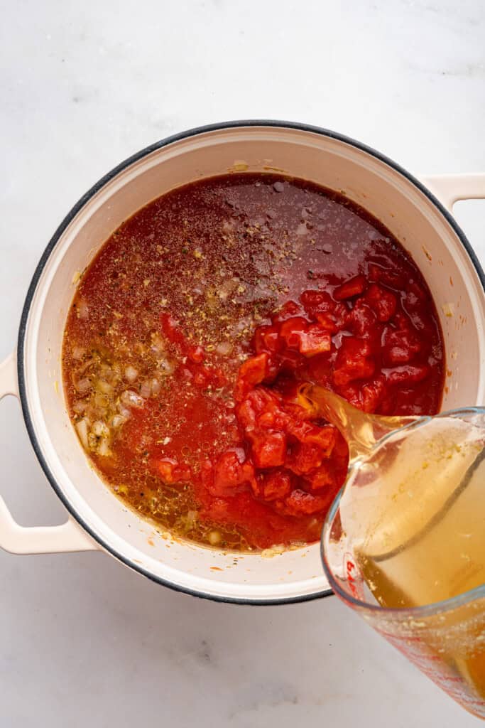 Pouring broth into a dutch oven with tomatoes and aromatics.
