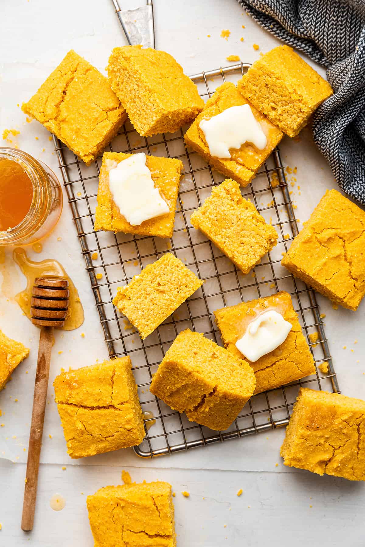 Slices of vegan pumpkin cornbread on a cooling rack, some with pats of vegan butter on them, with a jar of honey and a honey dipper sitting on the table