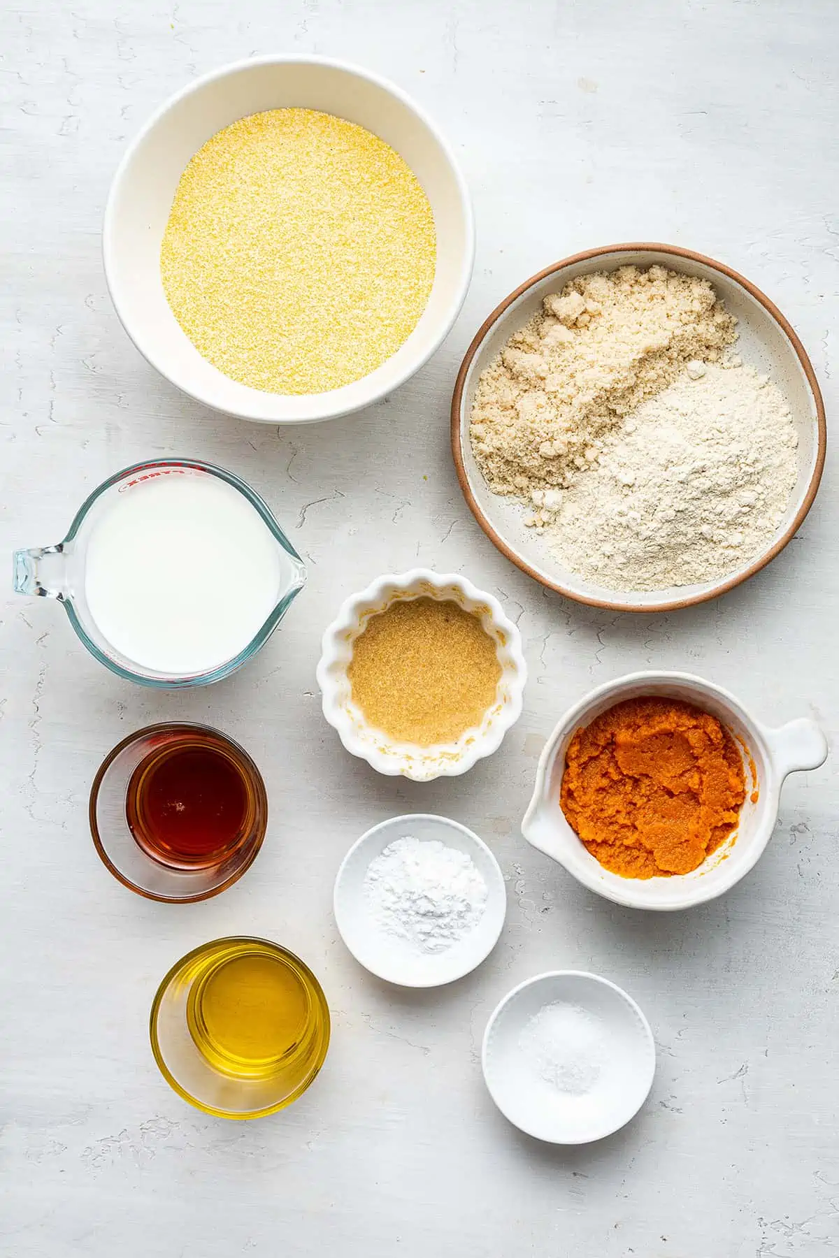 Overhead view of the ingredients for vegan pumpkin cornbread: cornmeal, almond flour, quinoa flour, pumpkin puree, a flax bomb, almond milk, salt, olive oil, maple syrup, and baking powder