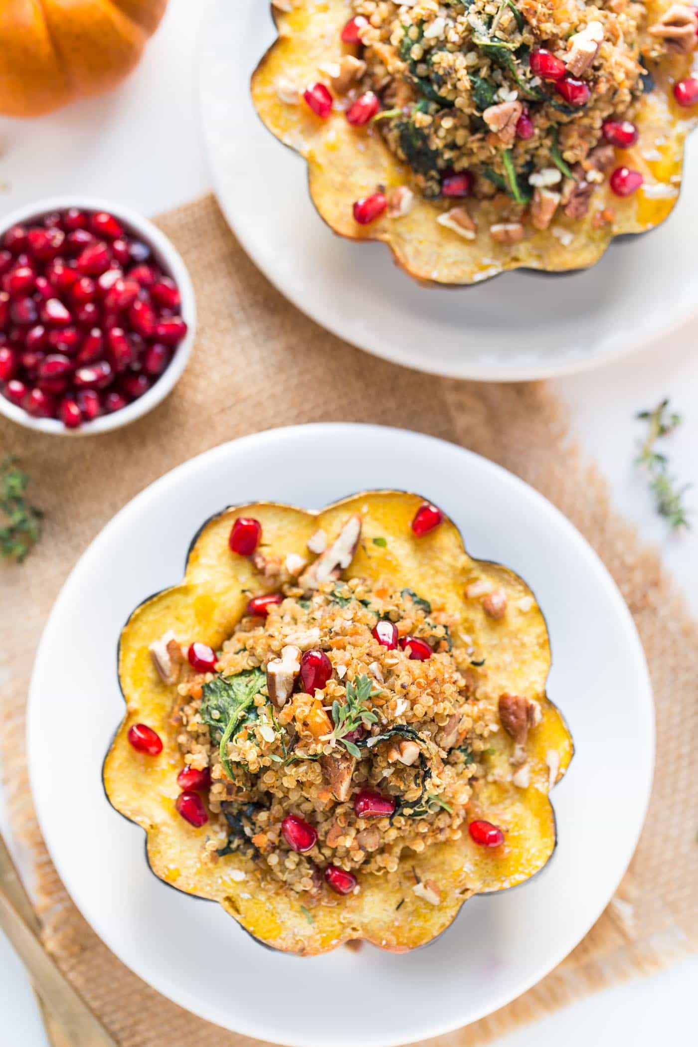 Overhead view of a mushroom stuffed acorn squash on a white plate, topped with pomegranate seeds, next to another plate with a serving of stuffed squash, and a bowl of pomegranate seeds