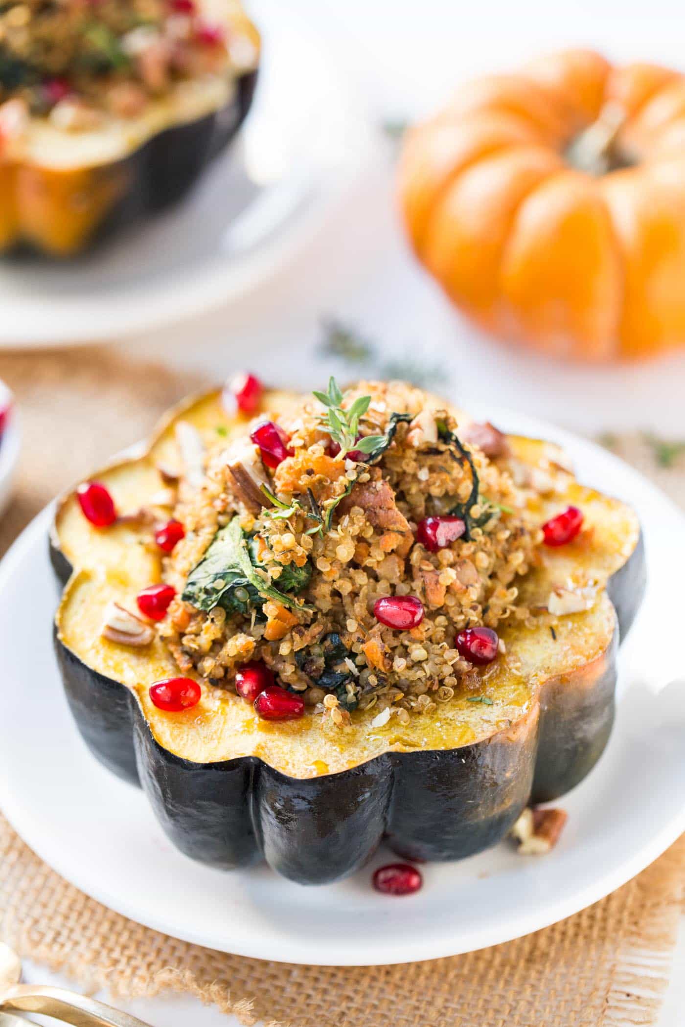 A mushroom stuffed acorn squash on a plate, topped with pomegranate seeds, with an uncooked acorn squash in the background