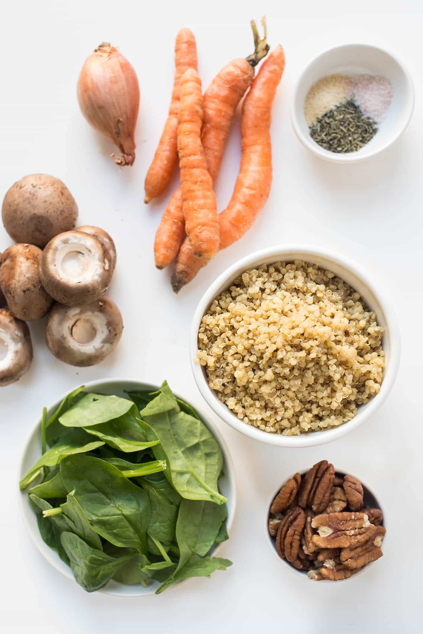 Overhead view of raw carrots, a shallot, raw baby bella mushrooms, a bowl of raw spinach, a bowl of pecans, a bowl of cooked quinoa, and a bowl of spices