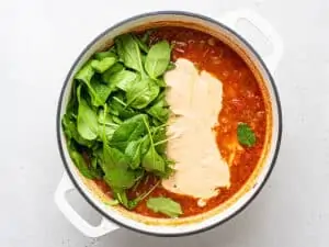 adding cashew cream and spinach to a pot of tomato soup