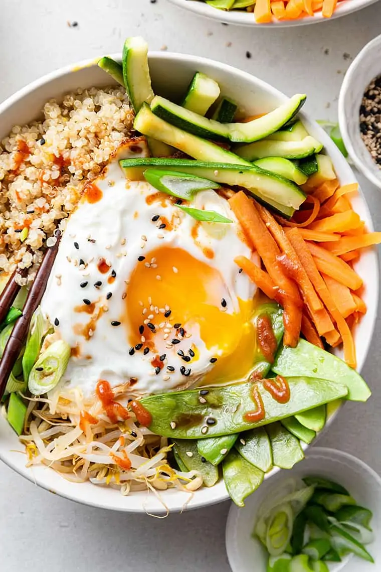 Closeup of quinoa bibimbap in white bowl with steamed veggies and runny sunny side up egg