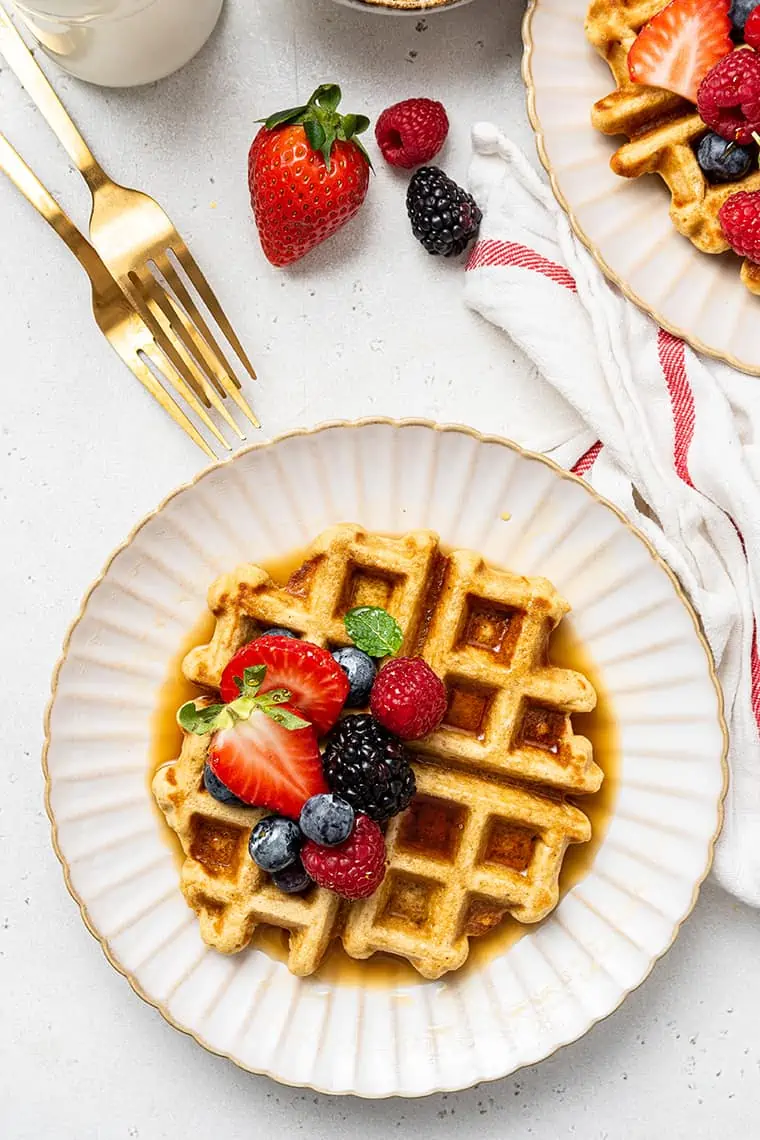 Top view of a waffle on a white plate with strawberries and blueberries, next to forks and another plate of waffles.