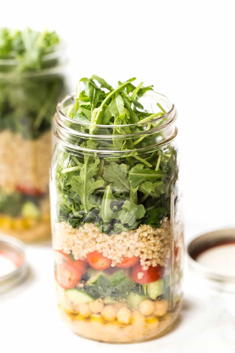 A mason jar overflowing with salad green. It also has quinoa, vegetables and chickpeas. Salad dressing fills the bottom. 