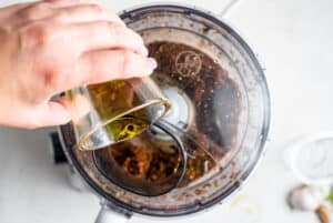 overhead of adding olive oil into a food processor making pesto