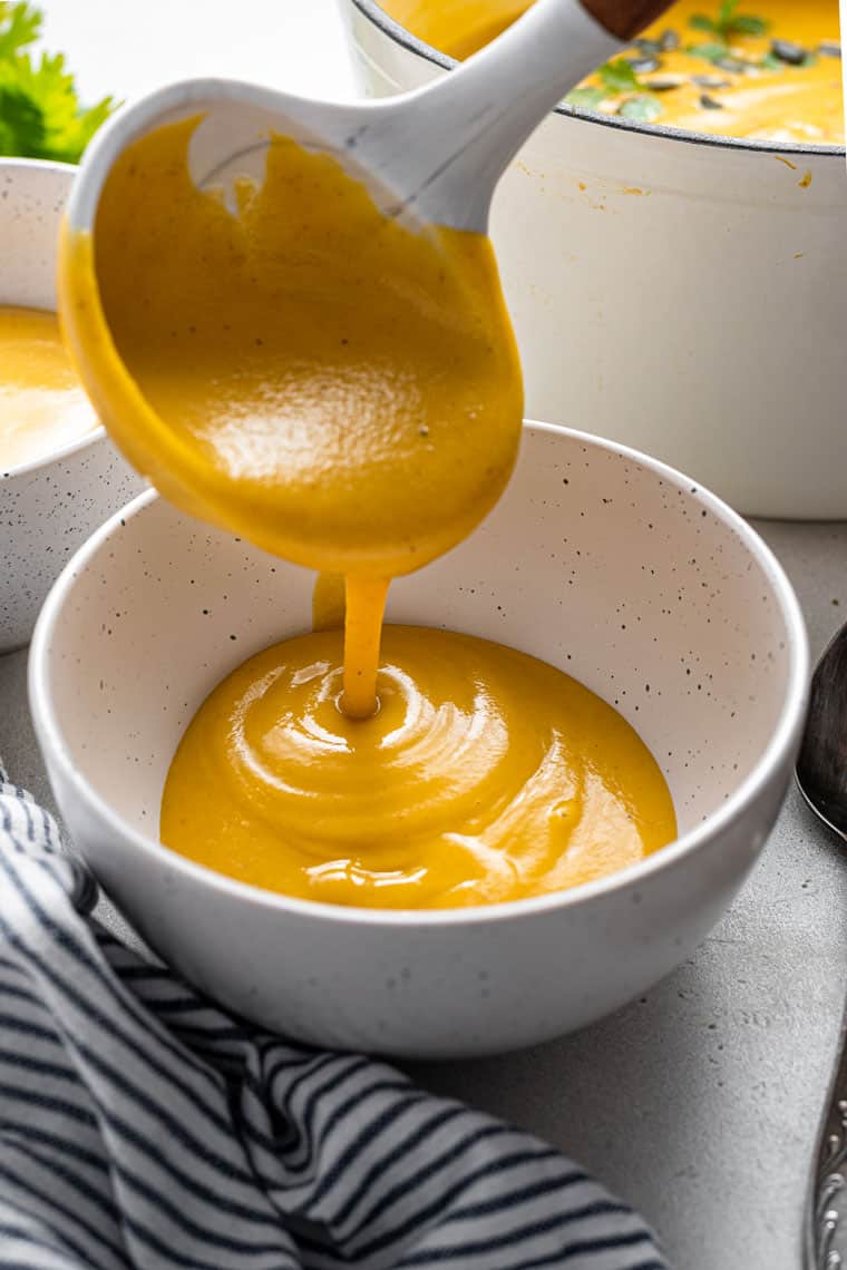 This curried acorn squash soup recipe is cozy and creamy, packed with roasted cauliflower, veggies, and spices. Vegan and freezer-friendly! pouring cauliflower and acorn squash soup into a bowl with a ladle