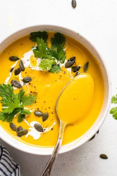 Overhead view of a bowl of acorn squash soup topped with a drizzle of cream, fresh cilantro, and pumpkin seeds with a spoon.