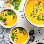 Overhead view of two bowls of acorn squash soup topped with cream, fresh cilantro, and pumpkin seeds, next to more soup in a large pot.