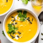 Overhead view of a bowl of acorn squash soup topped with a drizzle of cream, fresh cilantro, and pumpkin seeds next to a spoon.