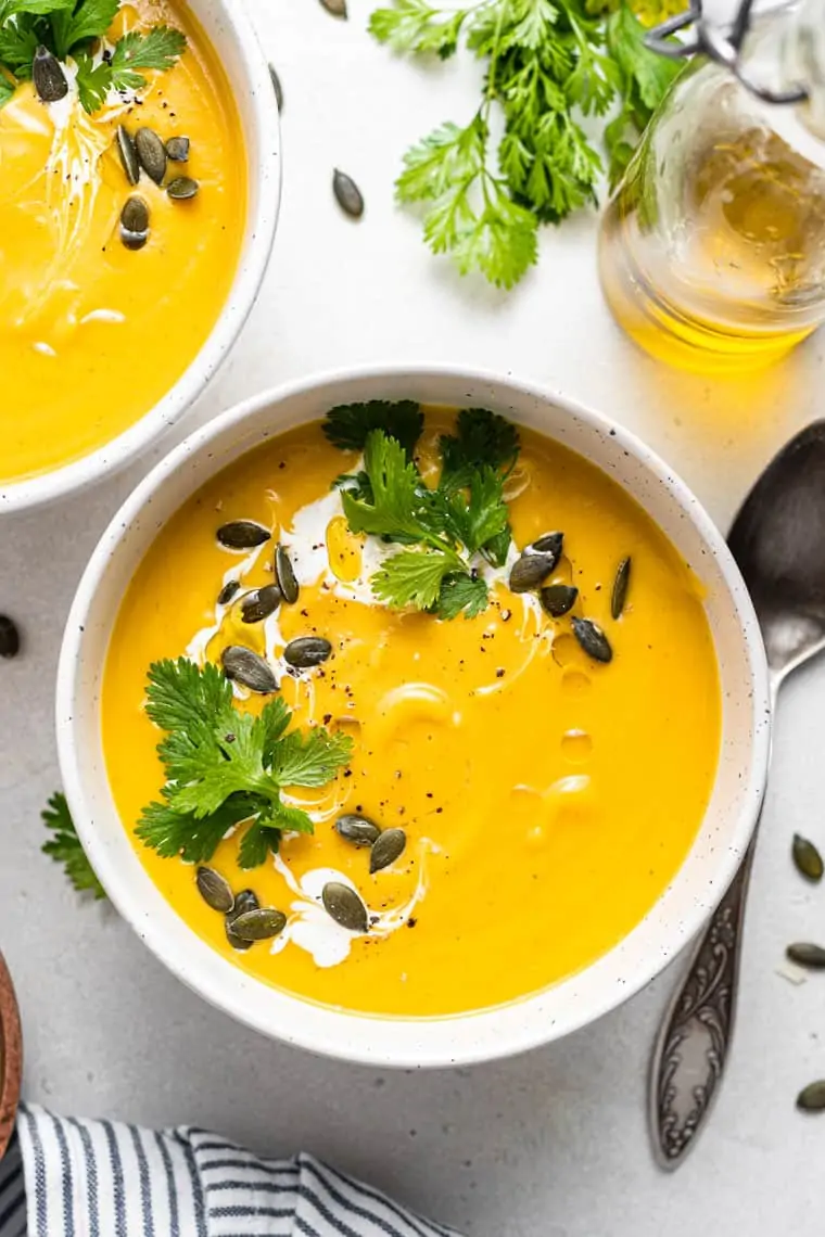 Overhead view of a bowl of acorn squash soup topped with a drizzle of cream, fresh cilantro, and pumpkin seeds next to a spoon.