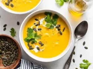 Overhead view of a bowl of acorn squash soup topped with a drizzle of cream, fresh cilantro, and pumpkin seeds next to a spoon.