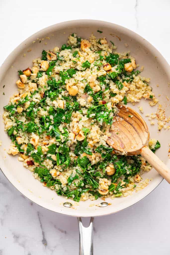 These Stuffed Sweet Potatoes are filled with quinoa, kale and hazelnuts, then topped with creamy tahini sauce. A hearty main or side dish! Quinoa, hazelnut, and kale mixture in skillet with wooden spoon.