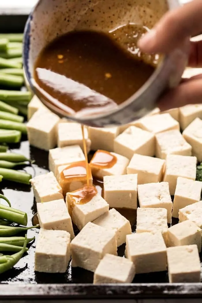 A bowl of sauce being poured over raw tofu cubes on a baking sheet