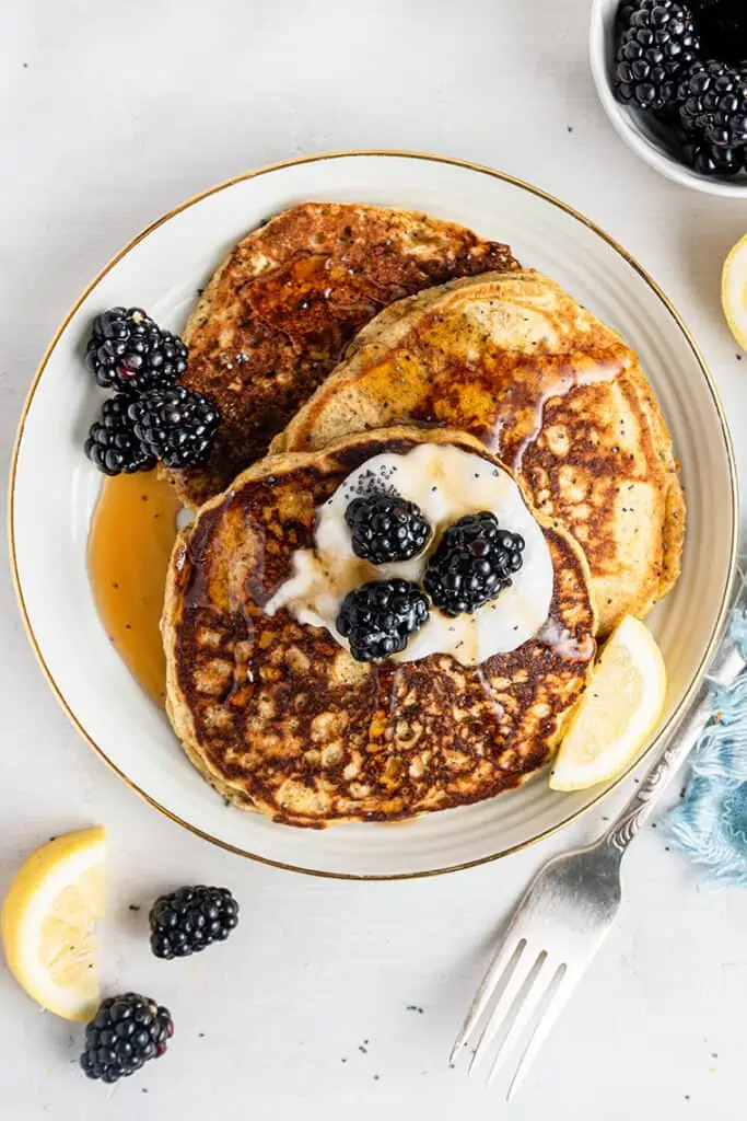 White plate with three lemon poppy seed pancakes topped with yogurt, blackberries and maple syrup