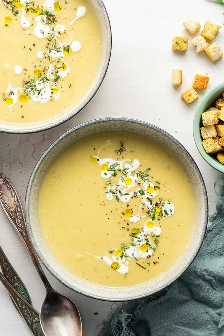 A bowl of vegan leek and potato soup, next to another bowl of soup and a bowl of croutons with the croutons spilling out