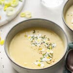 A bowl of vegan leek and potato soup garnished with vegan cream and rosemary, and in the background some raw leaks, rosemary, and croutons, and a glass