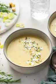 A bowl of vegan leek and potato soup garnished with vegan cream and rosemary, and in the background some raw leaks, rosemary, and croutons, and a glass