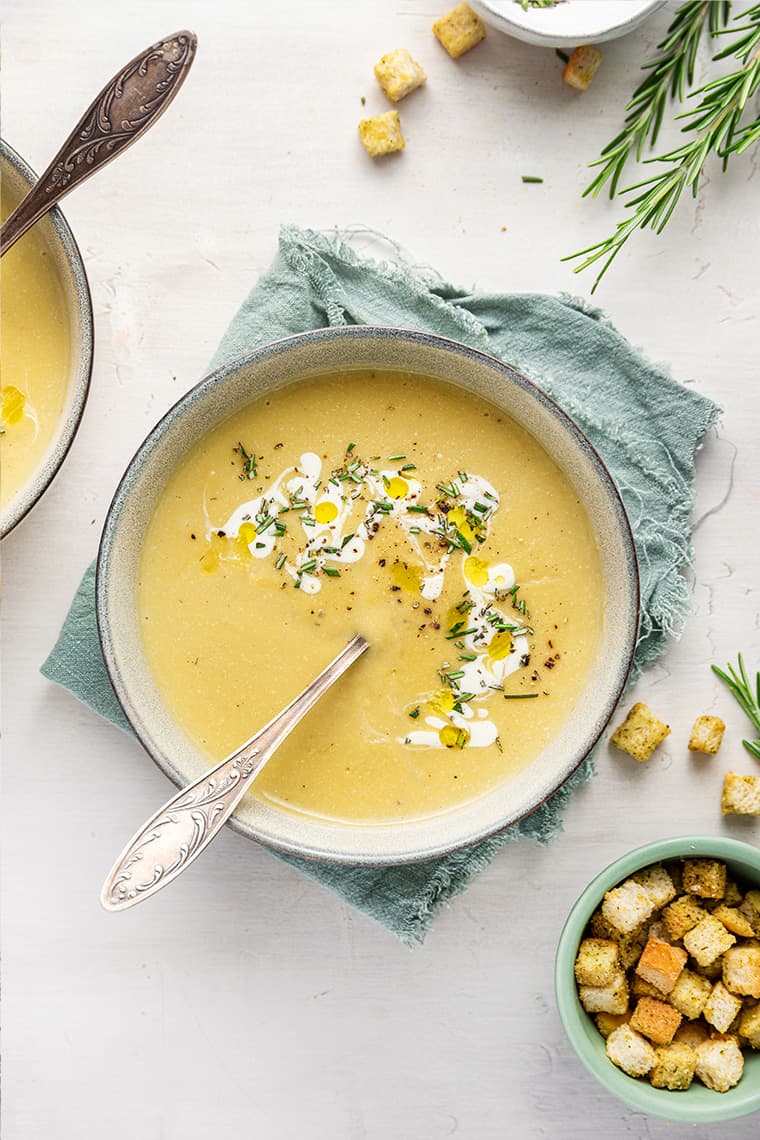 A bowl of vegan potato leek soup, garnished with vegan cream and rosemary, on a kitchen towel, with a spoon in the bowl, next to a bowl of croutons