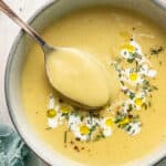 Close up of a bowl of vegan leek and potato soup, garnished with vegan cream and rosemary, with a spoon taking a spoonful out