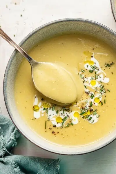 Close up of a bowl of vegan leek and potato soup, garnished with vegan cream and rosemary, with a spoon taking a spoonful out