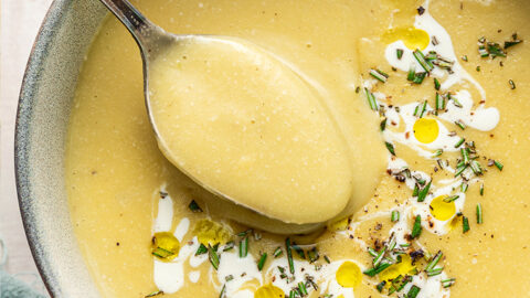 Close up of a bowl of vegan leek and potato soup, garnished with vegan cream and rosemary, with a spoon taking a spoonful out