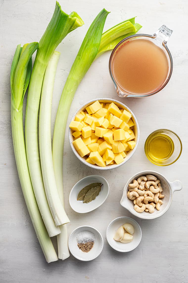 Overhead view of the ingredients needed for vegan leek and potato soup: raw leeks, cubed potatoes, vegetable broth, olive oil, raw cashews, salt, pepper, bay leaves, and garlic cloves