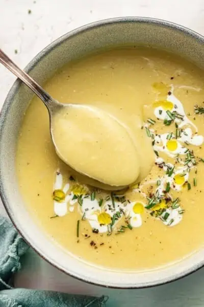 Close up of a bowl of vegan leek and potato soup, garnished with vegan cream and rosemary, with a spoon taking a spoonful out