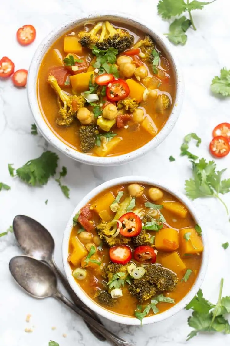 Overhead view of two bowls of vegetable curry, topped with chili slices and cilantro, surrounded by cilantro leaves, chili slices, and two spoons