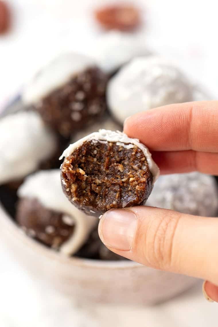 A guide for how to make energy balls, a protein and nutrient-rich snack full of nuts, seeds, and fruit, and easy to take on the go. Closeup of a hand holding an energy ball with a bite taken out of it, with a bowl of them in the background