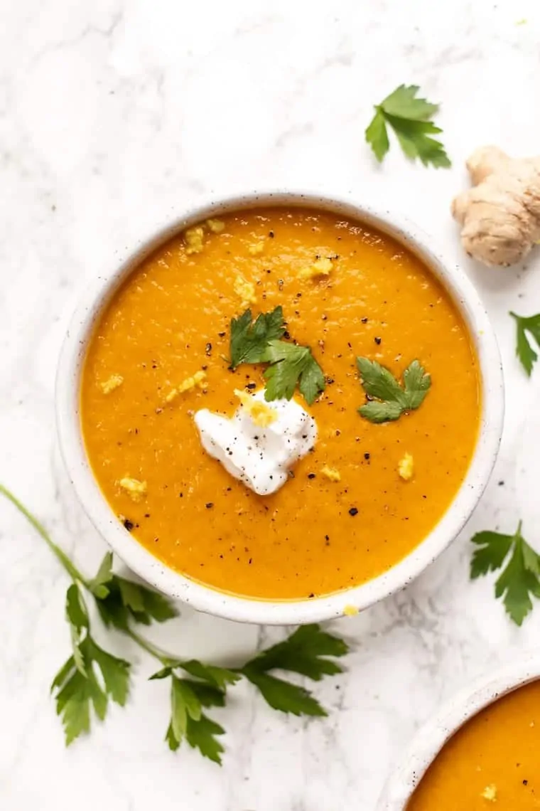 Overhead view of a bowl of turmeric carrot soup topped with vegan coconut yogurt, parsley, and cracked black pepper, surrounded by parsley, and whole ginger
