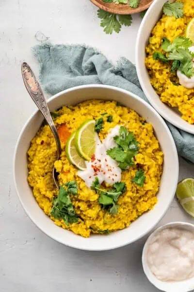 Overhead view of a bowl of kitchari topped with yogurt, cilantro, and lime slices, with a spoon in it, next to another bowl.